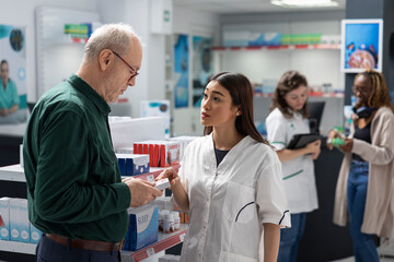 Asian pharmacist offers cardiology care advice to old man during pharmacy visit, recommending cardio medicine for heart health, cholesterol control and blood pressure prevention support.