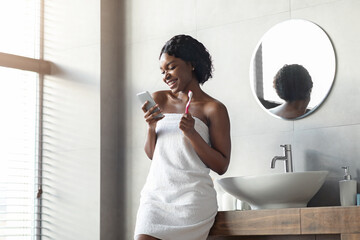 A cheerful young black woman, wrapped in a white towel, enjoys checking her smartphone while brushing her teeth. She stands in a modern bathroom with natural light.