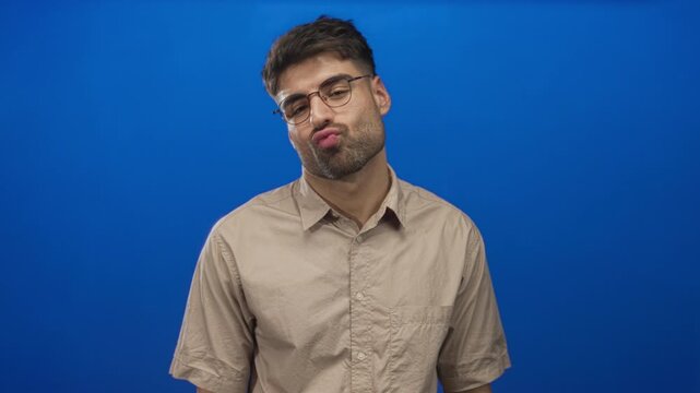 Man puckers lips and raises eyebrow wearing round glasses in studio against blue wall; playful flirtation.