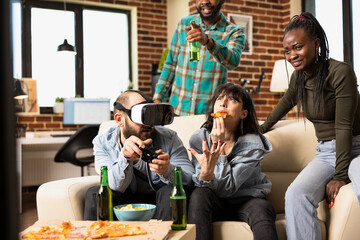 Caucasian male gamer playing virtual game tournament in cozy living room as friends eat pizza and observe game. White man with VR glasses and wireless controllers, having lively weekend at home.