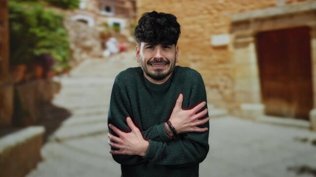 Young man shivering on an urban street, wearing a green sweater, expressing cold in a rustic town setting, showcasing discomfort on a chilly day outdoors.