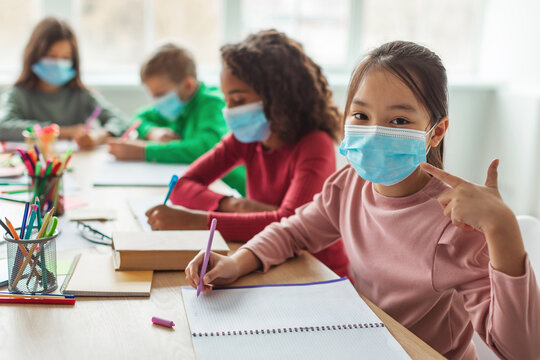In a modern classroom, a Japanese schoolgirl wearing a face mask smiles and gives a thumbs up.