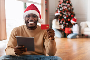 Fototapeta premium A happy man wearing a red Santa hat is sitting on the floor using a tablet and holding a credit card. In the background, his family is joyfully decorating a Christmas tree.