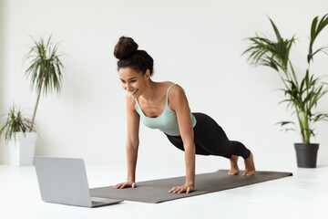 A sporty brunette woman stands in a plank position on a yoga mat while following an online workout tutorial on her laptop. She looks happy and focused during her ab workout.