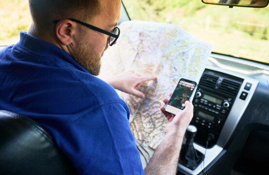 Man tourist sits in driver's seat of car, holding paper map and using smartphone with navigation app. Traveler finger points to location on map, cross-referencing routes to ensure accurate navigation.