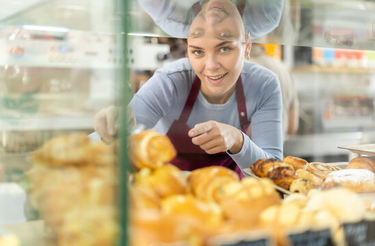 Positive young woman in apron offering fresh croissants standing behind glass counter in cozy cafeteria