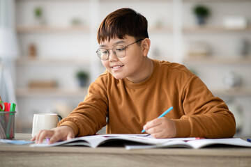 A cheerful asian schoolboy is happily writing in his notebook while doing homework at a wooden table.