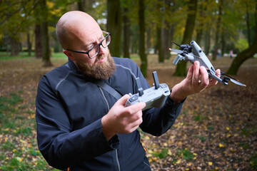 A man with a beard closely examines his drone while holding the remote control in a lush, colorful park during the autumn season.