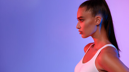 A sporty lady poses confidently, looking aside in a side profile. She wears white fitwear and stands before a dynamic pink and blue neon studio background, capturing a moment of athletic elegance. © Prostock-studio