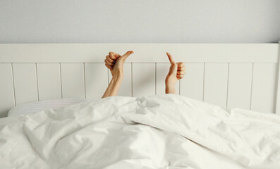 Cheerful woman waking up after sleeping in morning while lying in white bed from under the blanket
