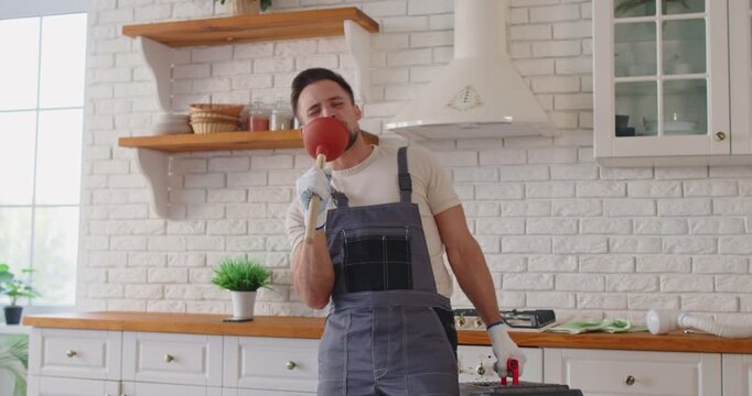 Plumber man singing, dancing with plunger in kitchen. Happy worker in uniform mimes into plunger, wearing gloves and smiling during fun break. Repair service and plumbing maintenance concept.