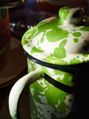 Closed view of a vintage-style green and white enamel teapot, featuring a splattered paint pattern and curved handle. This stylish kitchenware design is placed in a room with dramatic lighting. It's p