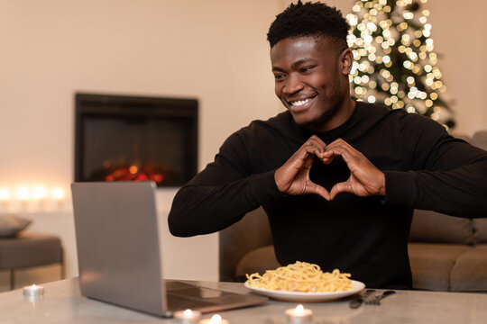 An African man is video calling and making a heart shape with his hands while enjoying Christmas dinner at home. A festive atmosphere is created by the Christmas tree in the background.