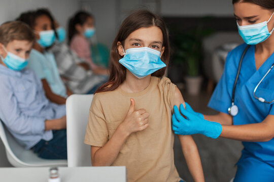 Girl wearing a face mask smiles and gives a thumbs up as a nurse places a bandage on her arm. Other children wait in the background at a vaccination clinic during coronavirus immunization.