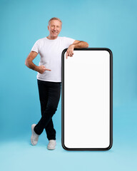 A mature man stands on a blue backdrop, leaning casually while pointing at a big empty smartphone...