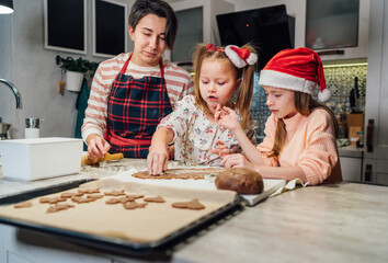 Cute little girls in red Santa hats with mother making homemade dough Christmas gingerbread cookies using cookie cutters together in home kitchen. Happy family holidays preparation, childhood concept.