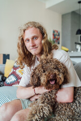 Portrait of young teenager boy with long curly hear smiling cheerfully sitting on comfortable living room sofa hugging Maltipoo dog showing joy of bonding with beloved pet