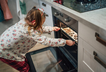 Little cute girl making homemade sweet biscuits, putting baking sheet into hot oven. Christmas cookies preparation, cozy home atmosphere, warm family life moments concept.