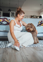 Middle aged female laughing cheerfully sitting on comfortable living room laminate floor hugging Maltipoo dog showing joy of bonding with beloved pet. Funny pets and heppiness concept.