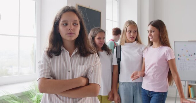 Bullying and baiting. Sad preteen girl is upset because of mockery of her classmates who openly mock her. Girl hides tears and covers face with hands when kids stand behind her and discussing her.