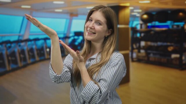 Young blonde woman presenting hands and smiling in bright indoor gymnasium with rows of cardio machines; happiness.