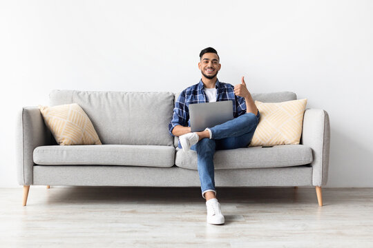 Happy Middle Eastern man enjoying a remote work session on his laptop while resting on a cozy couch. He shows a thumbs up sign, expressing satisfaction in his living room.