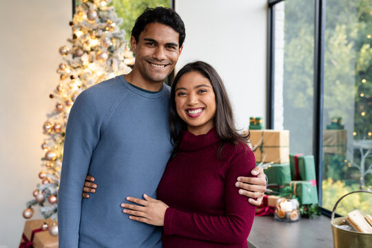 Asian couple smiling, standing arm in arm beside decorated Christmas tree with gifts in living room