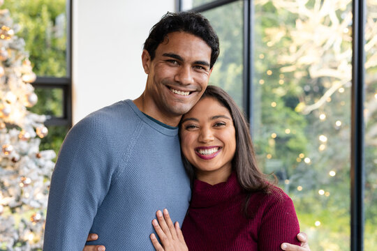 Couple standing by living room Christmas tree with string lights, metal ornaments and windows