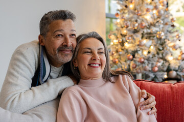 Diverse senior couple sitting on sofa in living room with Christmas tree lights and ornaments