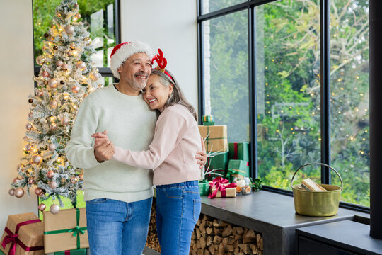 Diverse senior couple dancing at home by Christmas tree wearing Santa hat and reindeer headband