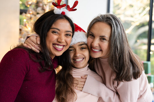 Three-generation family hugging in living room while wearing festive headgear beside Christmas tree