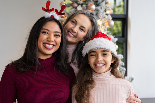 Diverse female family wearing antler headband and Santa hat standing by Christmas tree near window
