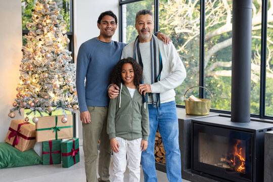 Three-generation male family standing at home with snow-frosted tree, wrapped gifts and wood stove