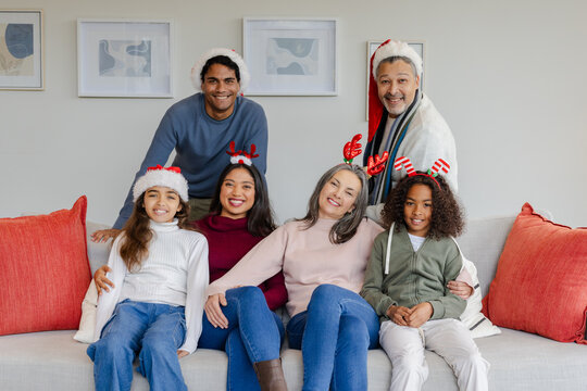 Diverse three-generation family sitting on sofa in living room wearing festive headwear