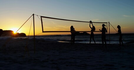Playing five women wearing swimwear and cap spiking volleyball on beach at sunset, with net