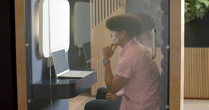 Sitting man in short-sleeve pink shirt leaning over laptop in glass pod, with slat wall - Powered by Adobe