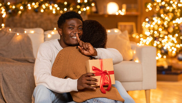 In a warm living room adorned with Christmas lights, a young black couple enjoys a heartfelt moment. The husband holds a gift box while signaling for silence, creating a special surprise for his wife. - Powered by Adobe