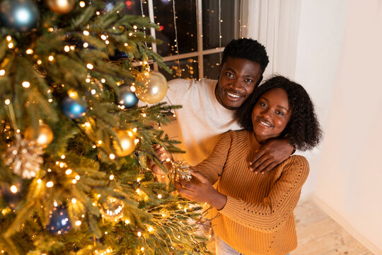 A joyful millennial black couple is decorating their Christmas tree with lights and ornaments in a warm living room. They share love and happiness while preparing for the holidays together. - Powered by Adobe