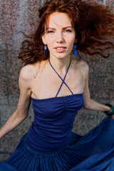 Fototapeta premium Confident Redhead Posing in Vibrant Blue Dress Against Industrial Urban Wall, Windblown Hair, Chic Fashion Shot