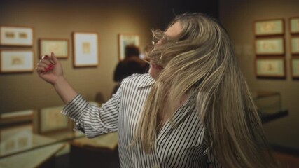 Woman flipping hair near framed artwork in gallery building hall, eyes closed and lips pressed; joy.