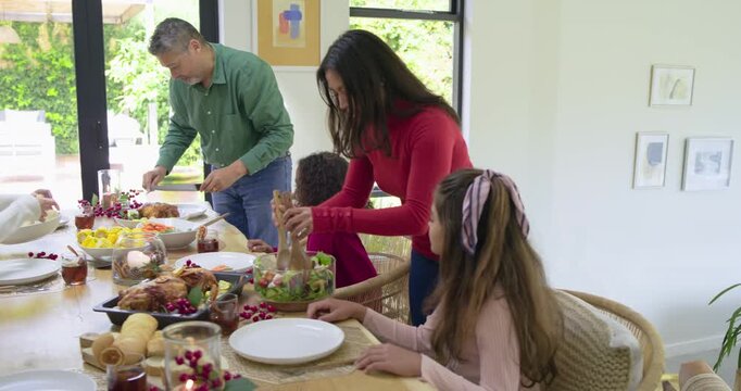 Diverse family starting holiday meal dad carving turkey and mom tossing salad at table serving kids