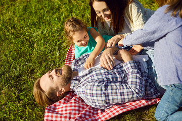 Happy family lying together on blanket outdoors, laughing, children tickling father, smiling in...