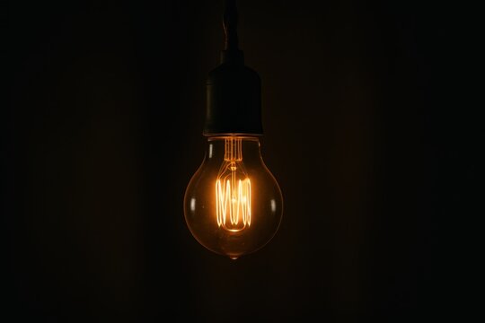 Single glowing glass light bulb hanging in moody dark room with soft warm bokeh
