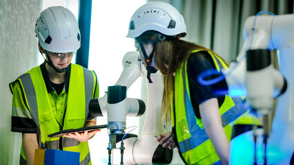 Two female engineers in hard hats and safety vests test a high tech robotic arm. One holds a...