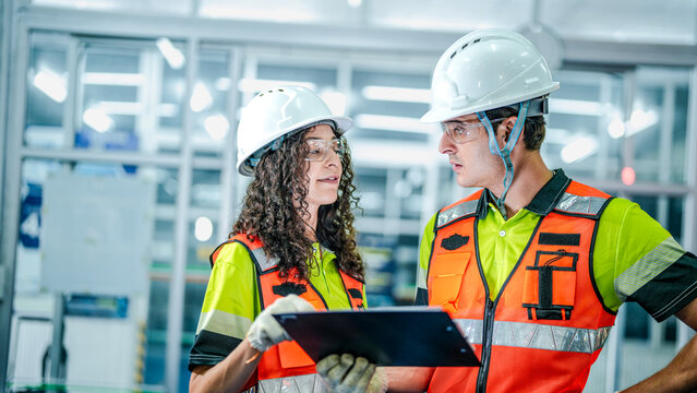 Two engineers use a tablet to program an AI system. They are in a futuristic smart factory, reviewing data for industrial robots or microchip manufacturing.