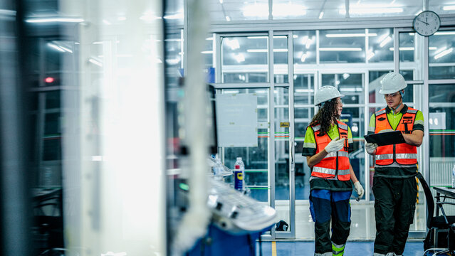 A male and female engineer in hard hats and safety vests use a tablet. They are collaborating in a modern, high-tech smart factory or cleanroom.