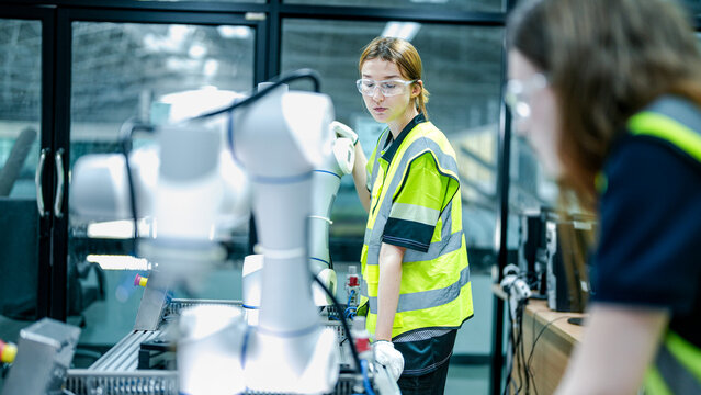 Two female engineers in safety vests collaborate in a tech lab. One woman in gloves inspects a high tech robotic arm while her colleague holds a tablet. - Powered by Adobe