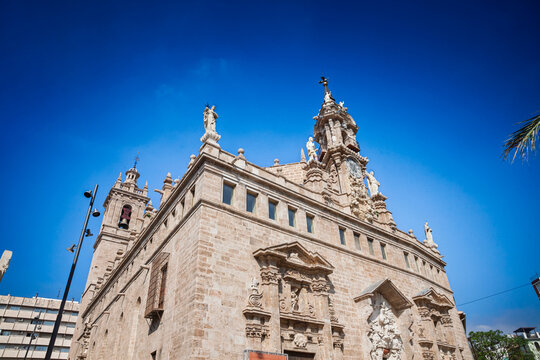 Iglesia de los Santos Juanes(sant juan del mercat) in Valencia with its Baroque facade, clock and rooftop statues. The stone exterior and sculpted portal fill the frame under a bright blue sky.