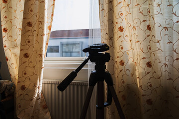 A dark camera tripod is silhouetted against a bright window framed by sheer, beige curtains patterned with floral vines.
