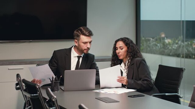 Two professionals engage in a focused conversation at a modern corporate office. They review documents and analyze data on a laptop, demonstrating teamwork and collaboration. - Powered by Adobe